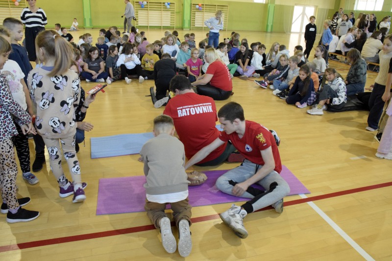 Grupa uczniów siedzi dokoła, w środku młodzi ratownicy pokazują dzieciom, które klęczą nad manekinami , jak wykonać uciśniecia klatki