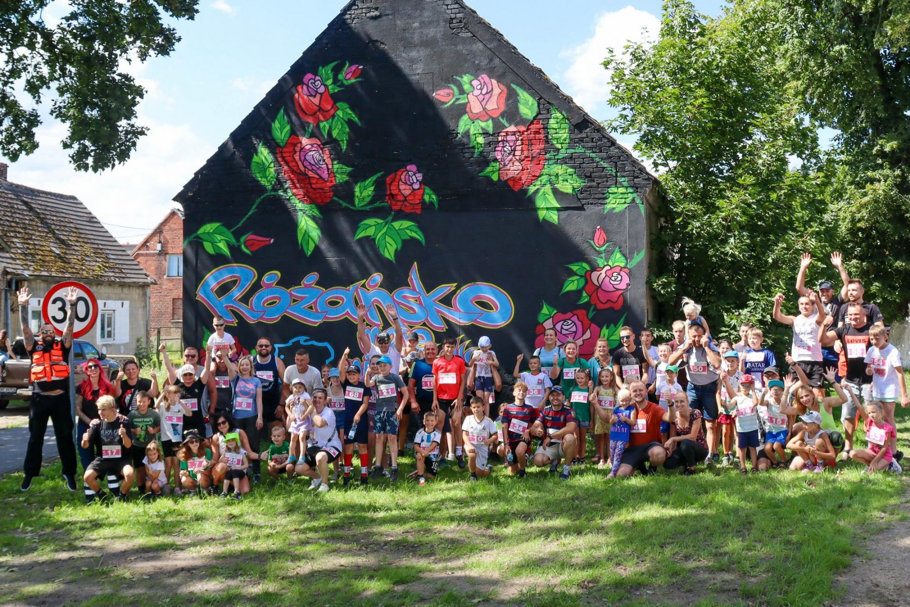 Grupa mieszkańców z numerami startowymi stoi na tle budynku z muralem 