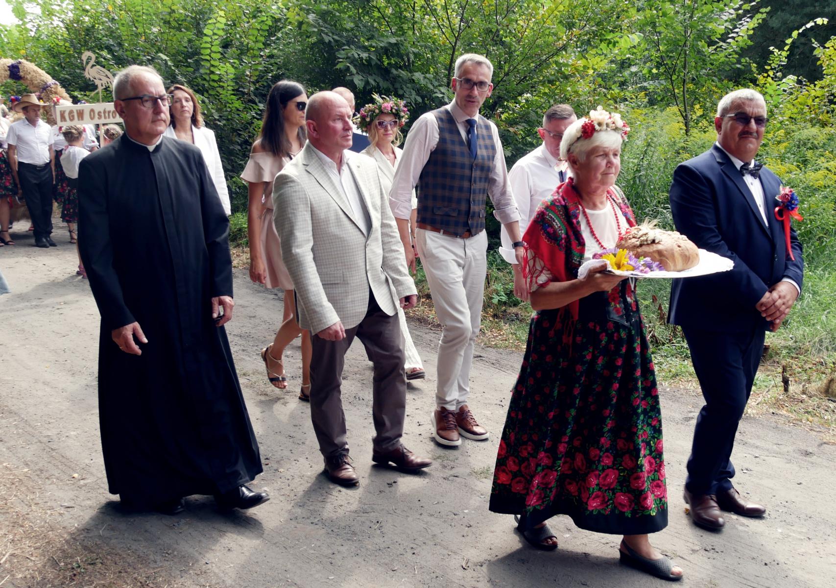 Korowód dożynkowy. w pierwszej lini starstowie z chlebem, w drugiej lini ksiądz, sołtys, burmistrz, przewdoniczący rady , za nimi inne osoby