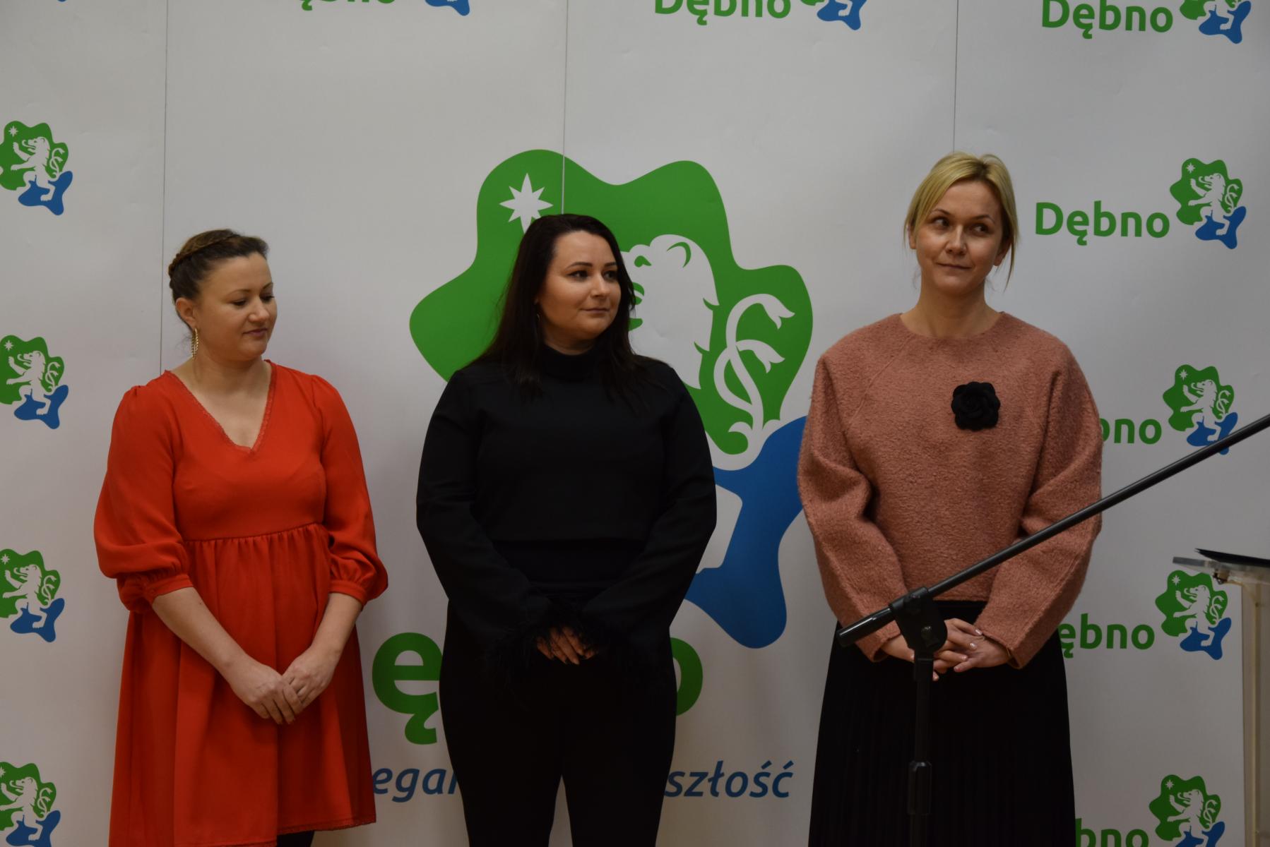 Trzy nauczycielki stoją, w tle biała ścianka, z zielono- niebieksim logiem i napisem Dębno. 