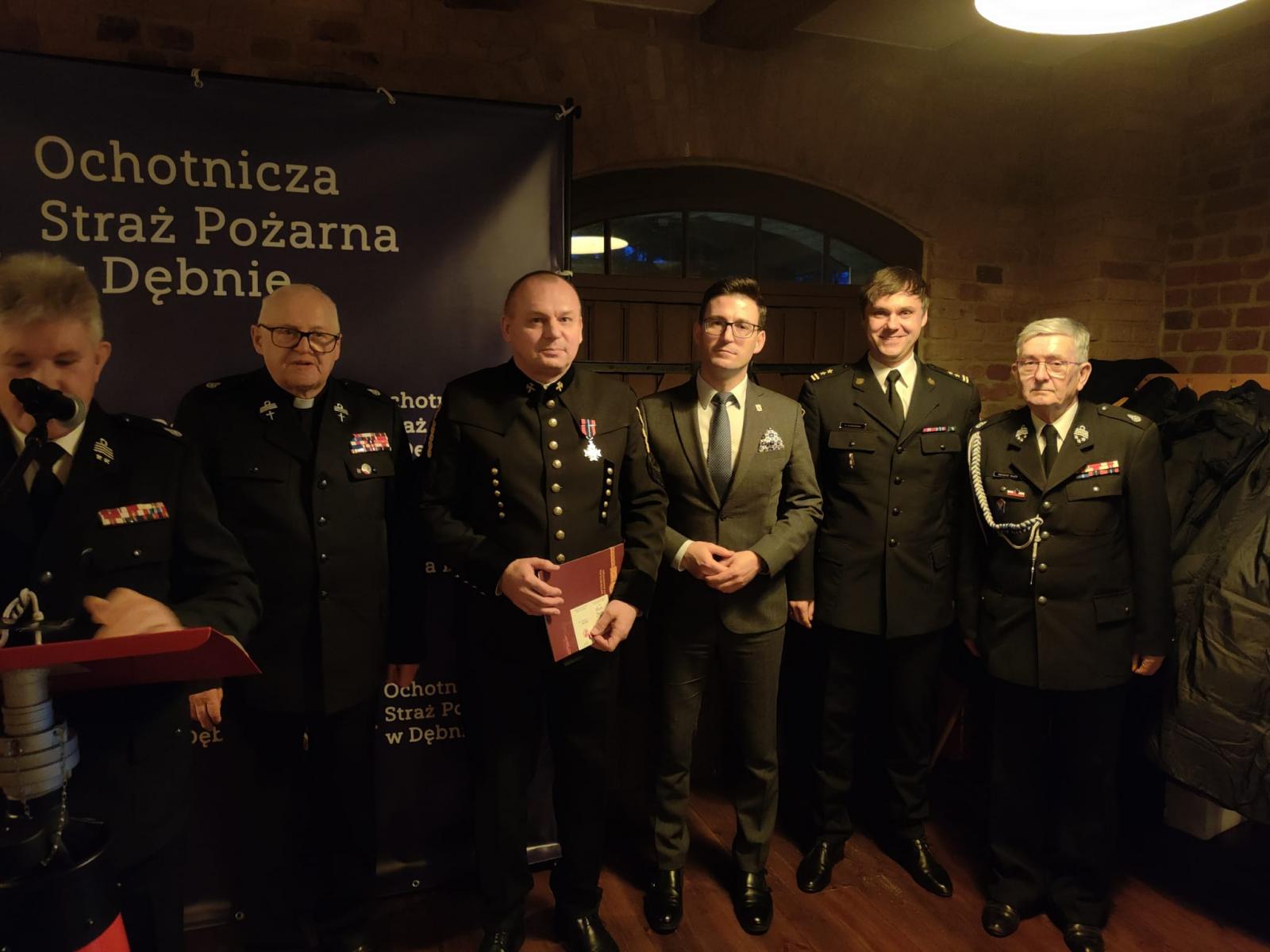 Przy mównicy stoi prezes OSP, za nim pieciu mężczyzn, zaproszeni goście 