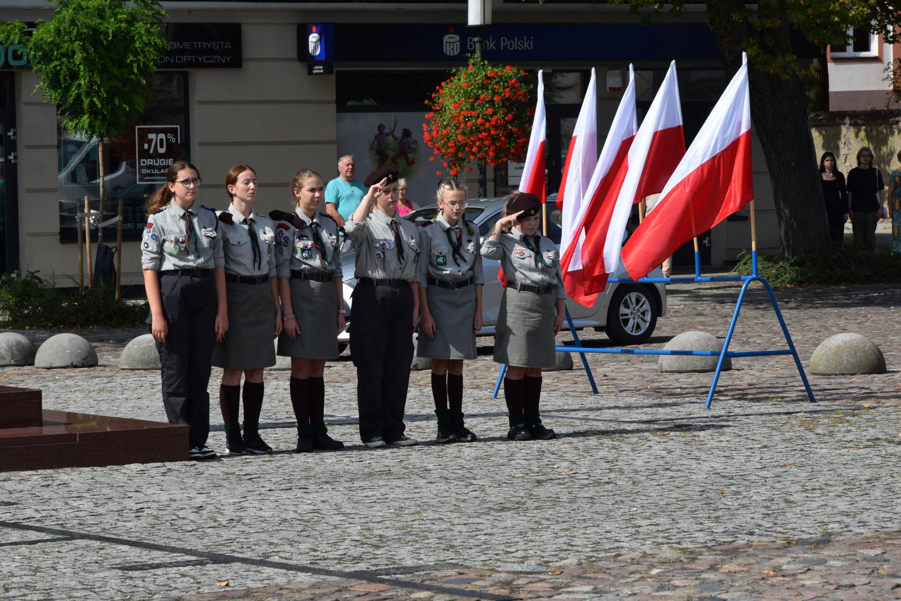 Harcerki stoją na placu. w tle powiewają biało-czerwone flagi