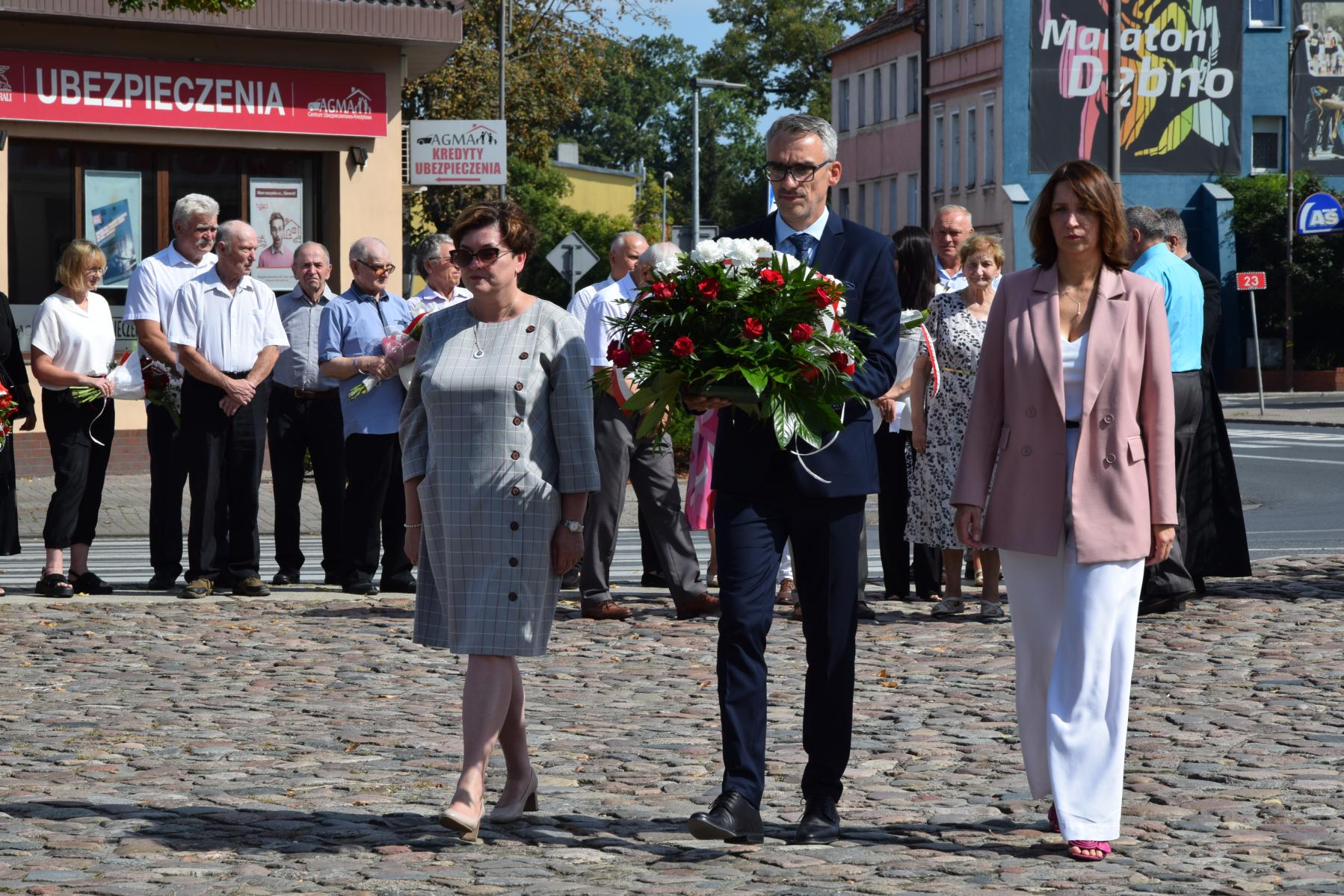Delegacja samorządu, burmistrz w środku między paniami, trzyma biało-czerwone kwiaty