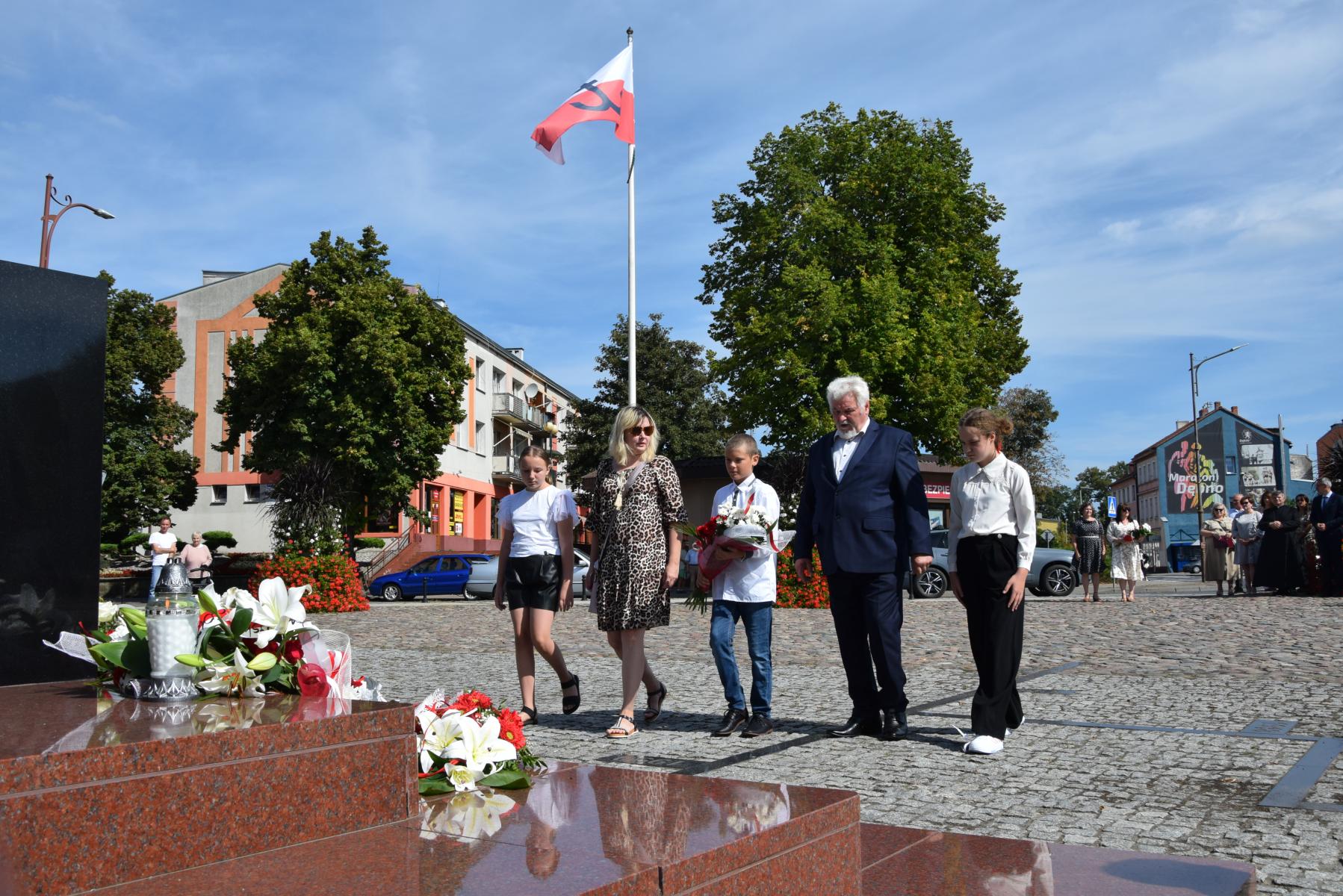 Dzieci z osobami dorosłymi, w tle powiewa biało-czerwona flaga 