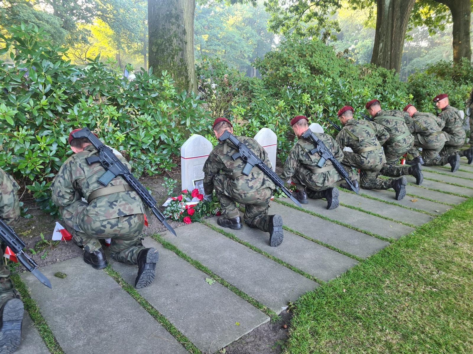 Żołnierze w szeregu klęczą przed nagrobkami, w tle drzewa zielone krzewy