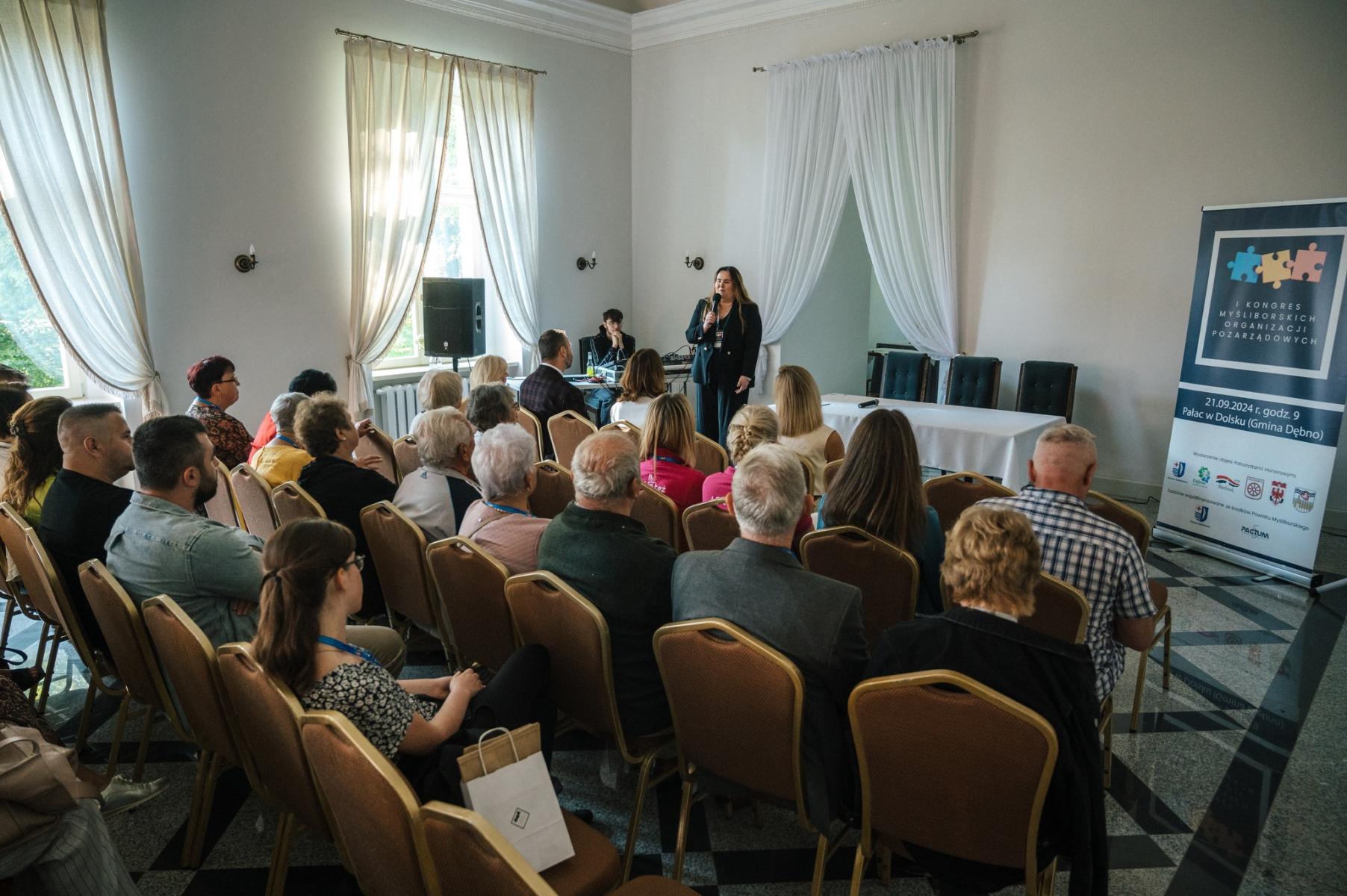 Ludzie siedzą w rżedach na krzesłach. Z przodu przemawia organizatorka. W sali białej