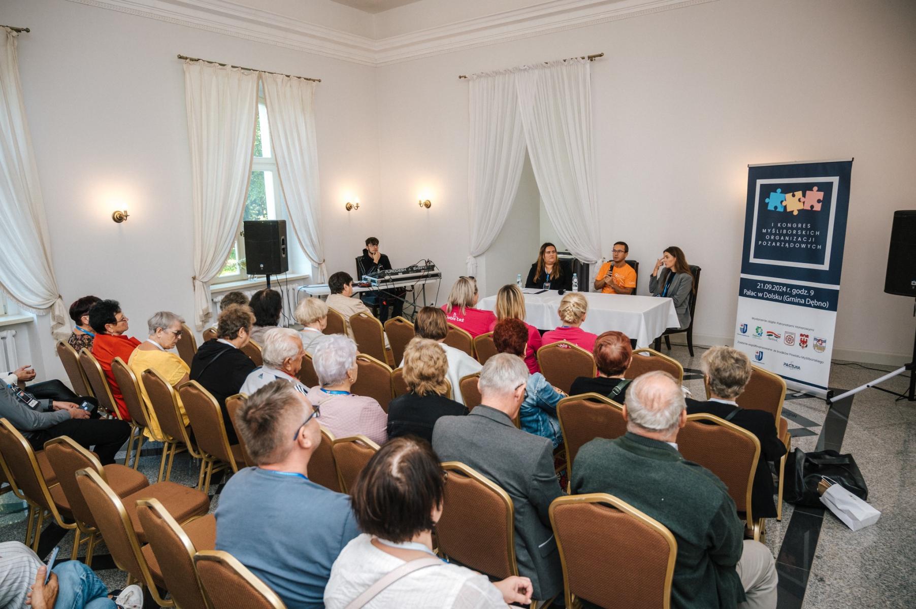 Uczestnicy panelu siedzą na krzesłach ustawionych w rzedach. Przed nimi przy stole siedzą trzy osoby prowadzące