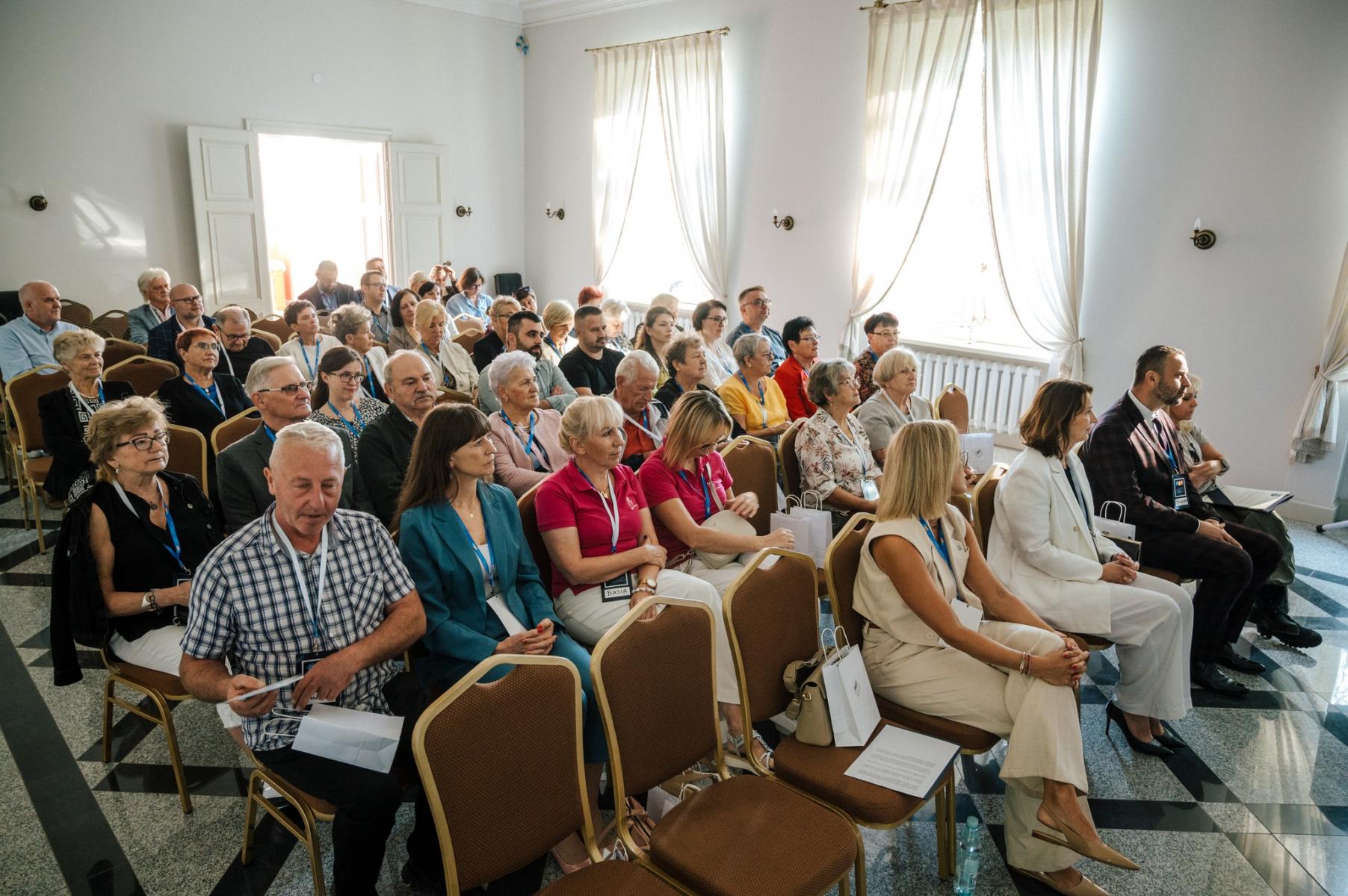 Uczestnicy Kongresu siedzą na krzesłach w rzędach. Wnętrze sala z białymi ścianami