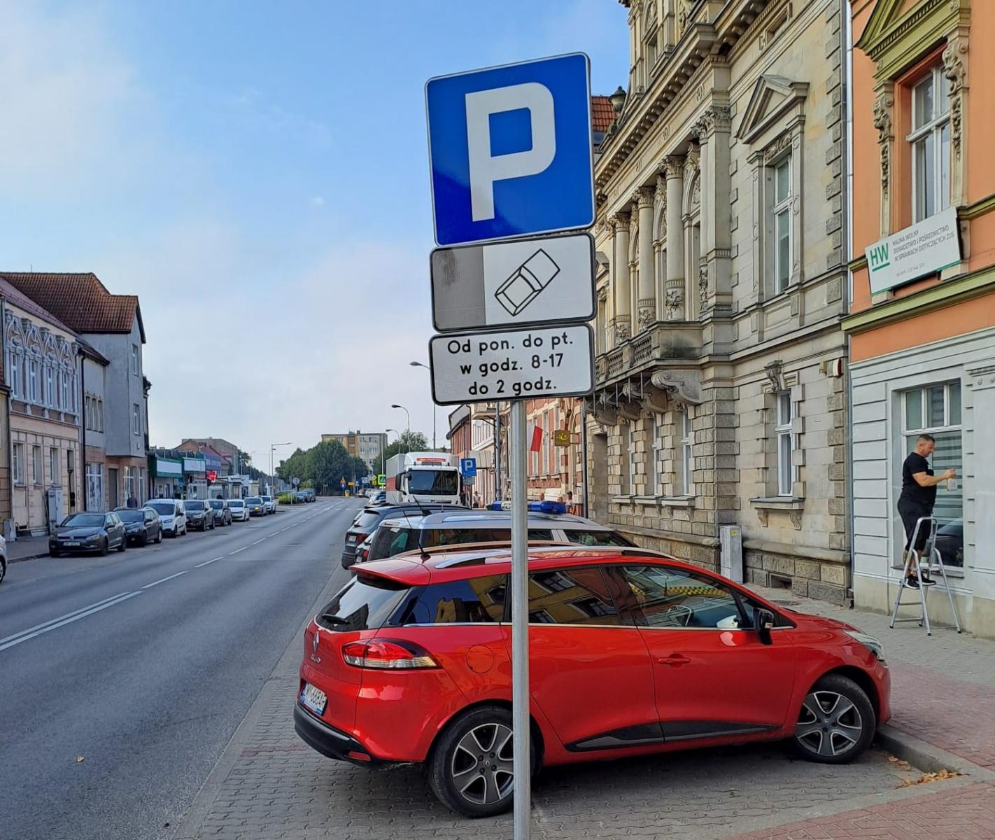 Rząd samochodów stoją na parkingu. Znak drogowy Parking z informacją jak i kiedy parkować. Po obu stronach budynki 