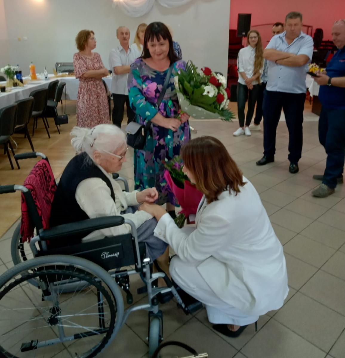Zastępca burmistrza w białym garniturze  kuca obok jubilatki, która siedzi na wózku. Z tyłu obok nich stoi sołtys wsi, trzyma w rękach kwiaty