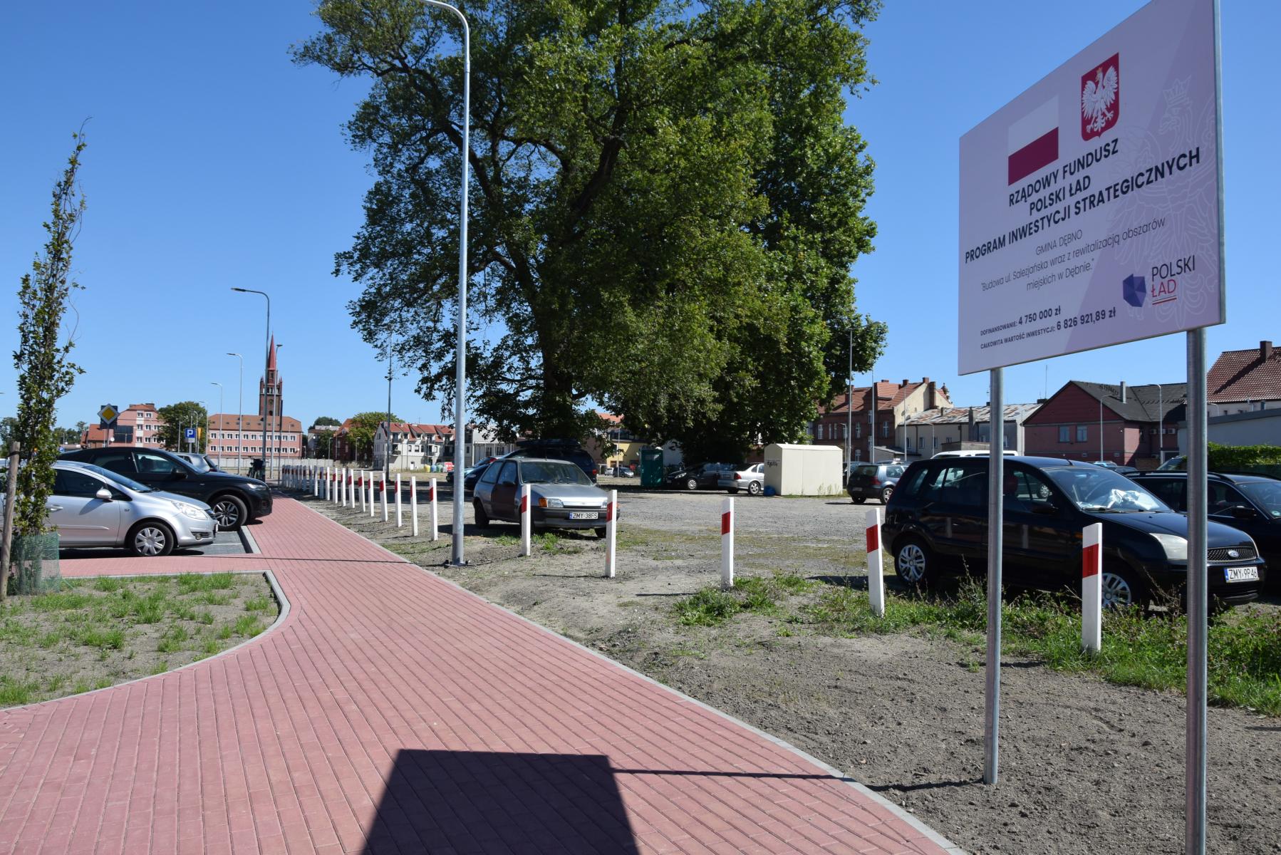 Chodnik, tablica z informacją o dofinansowaniu, drzewa, samochody na parkingu
