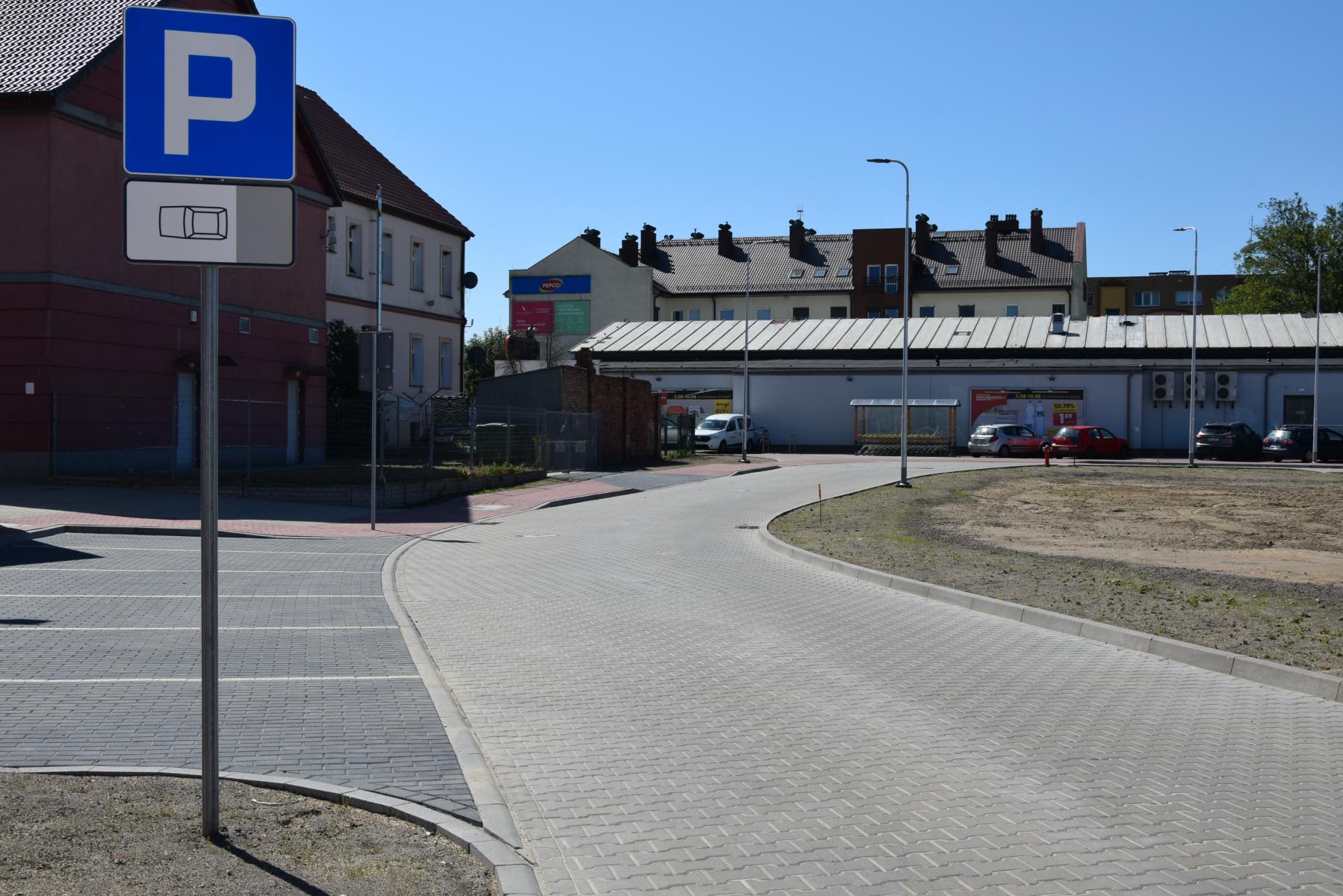 Droga, jezdnia z szarj kostki, znak Parking, miejsca parkingowe z kostki grafitowej, lapmy , w oddali budynki