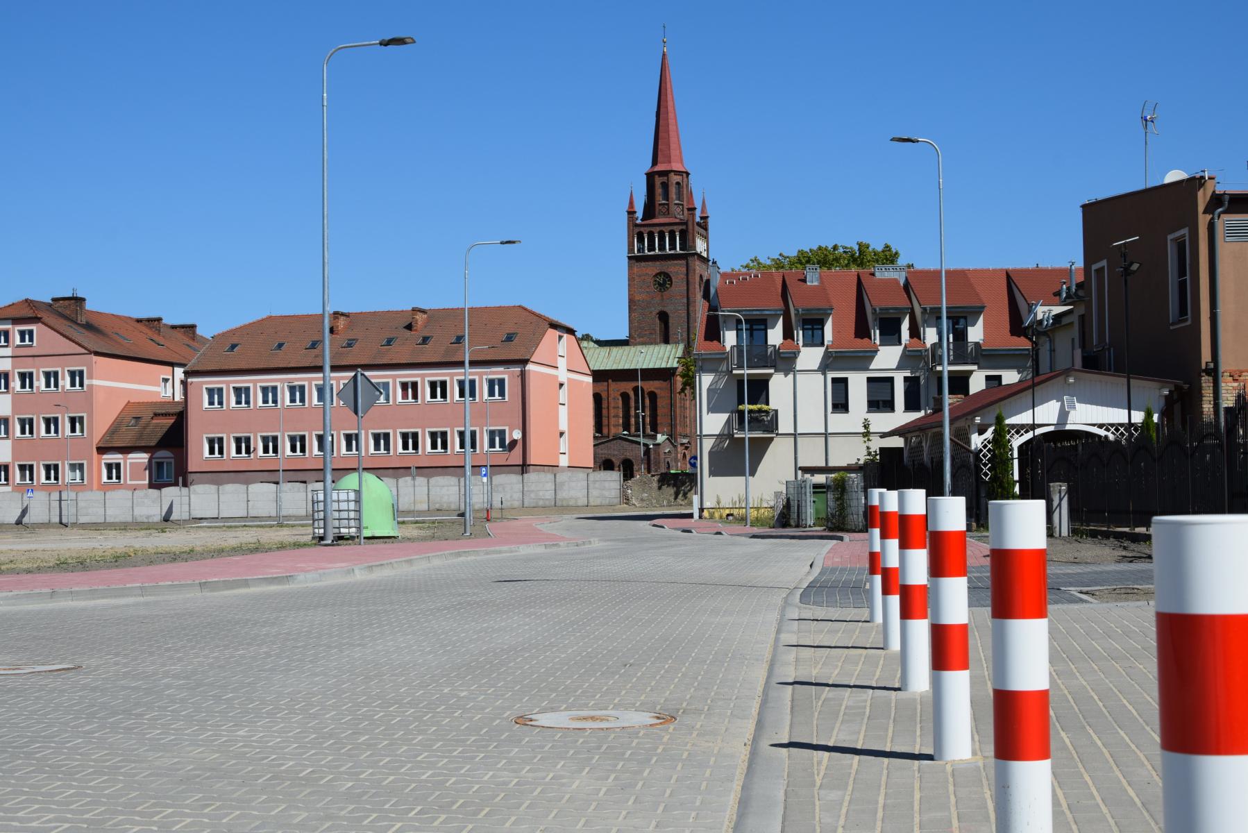Jezdnia z kostki szarej, po prawej rząd słupków biało czerownych, w odali budynki i wieża kościoła.