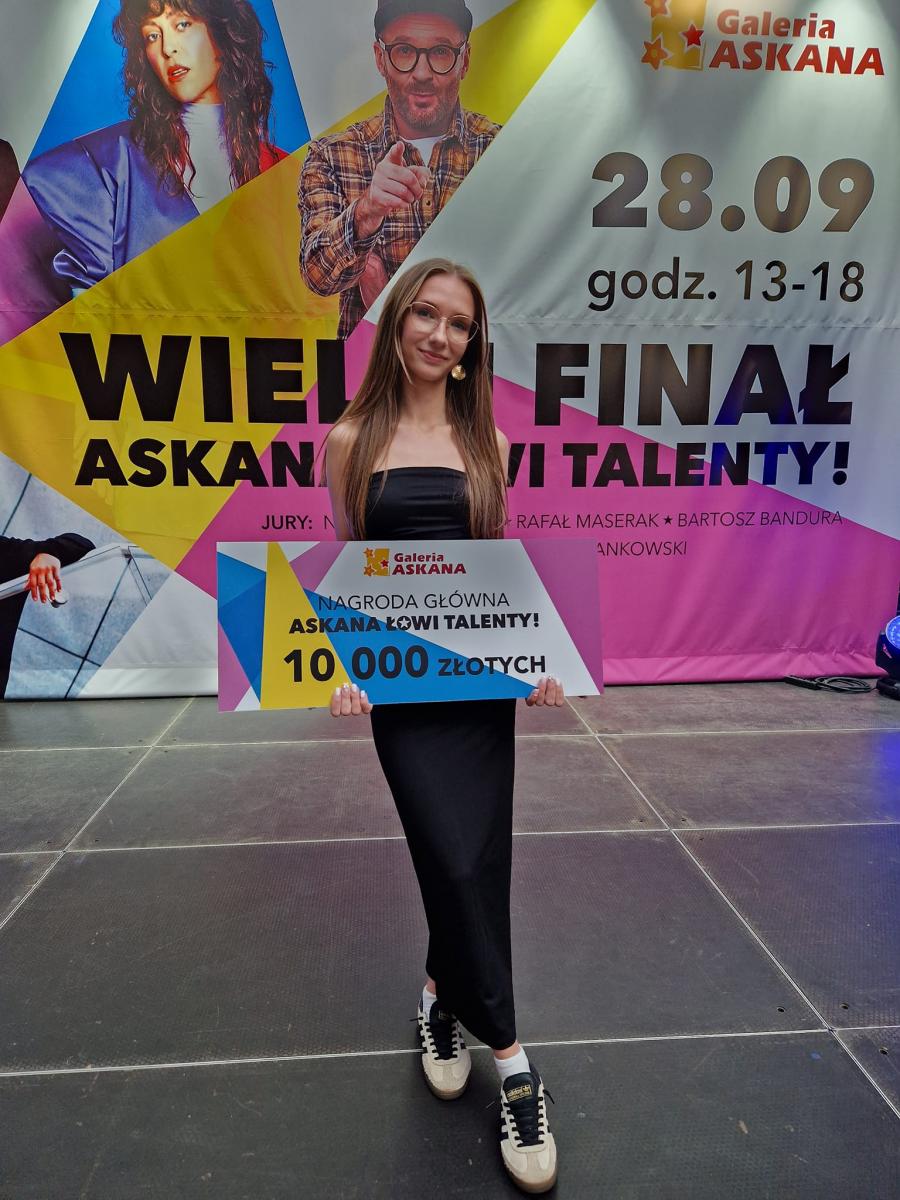 Powiększ zdjęcie. Wiktoria Taracińska stoi na scenie. trzyma czek na 10 ty złotych. W tle baner promujący wydrzenie 