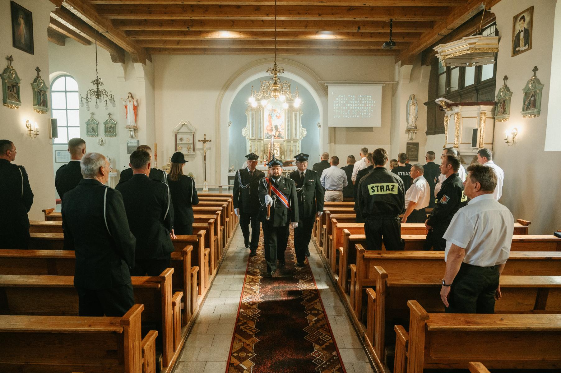 Strażacy w sztandarze idą przez środek kościoła, po bokach w ławkach stoją ludzie 