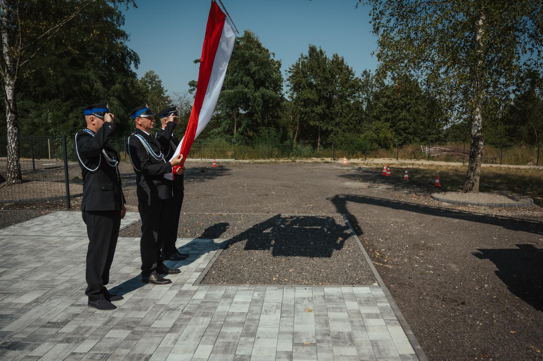 Trzech strażaków stoi, dwóch po bokach salutuje, środkowy trzyma biało-czerwoną flagę 