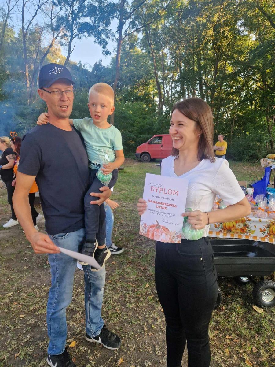 Uśmiechnięta kobieta trzyma dyplom w rękach. Obok stoi mężczyzna z chłopczykiem na rękach
