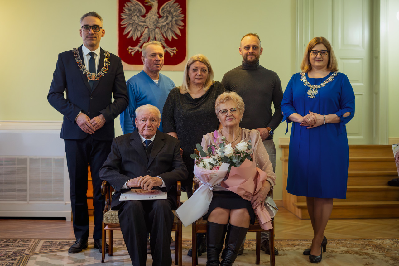 Para małżeńska siedzi na krzesłach, z tyłu stoją burmistrz i goście zaproszeni goście 