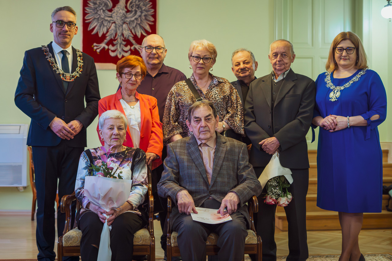 Para małżeńska siedzi na krzesłach, z tyłu stoją burmistrz i goście zaproszeni goście 