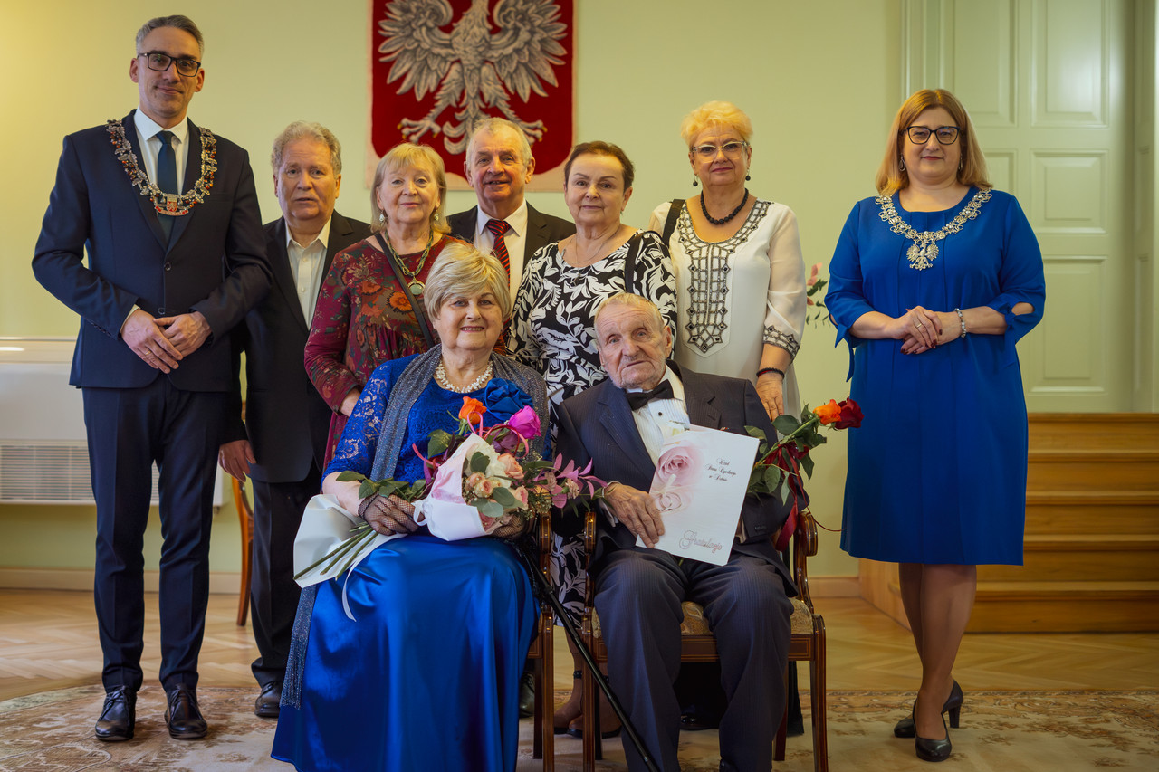 Para małżeńska siedzi na krzesłach, z tyłu stoją burmistrz i goście zaproszeni goście 