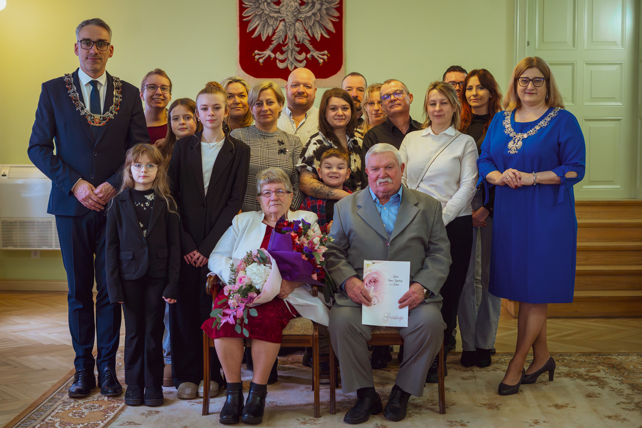 Para małżeńska siedzi na krzesłach, z tyłu stoją burmistrz i goście zaproszeni goście 