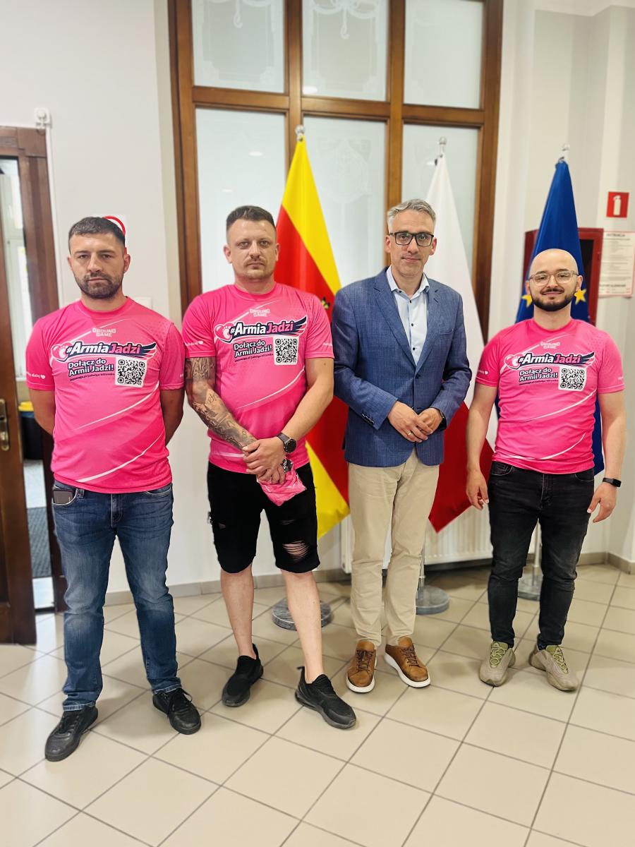 Organizatorzy stoją w różowych koszulkach, w środku stoi burmistrz w holu