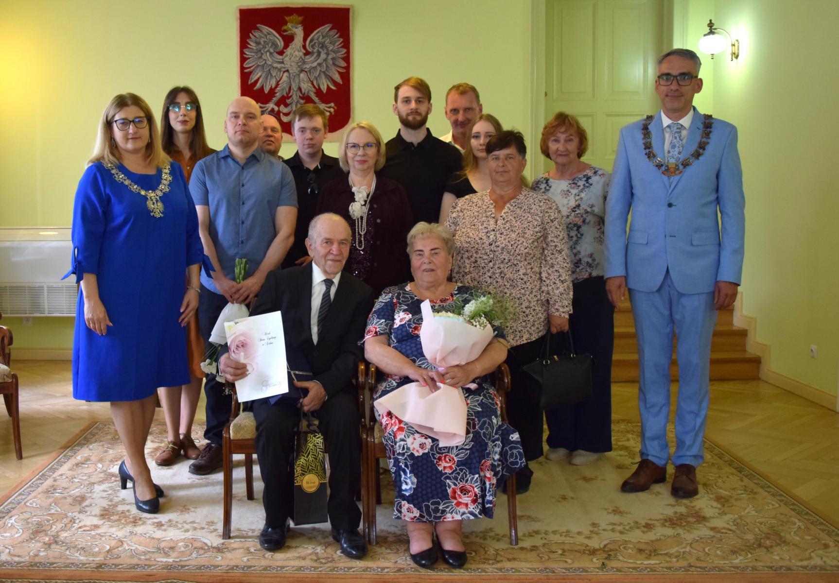 Jubilaci Państwo Majcherowie siedzą na krzesłach w sali, za nimi stoi rodzina, pani kierownik z USC i Burmistrz Dębna