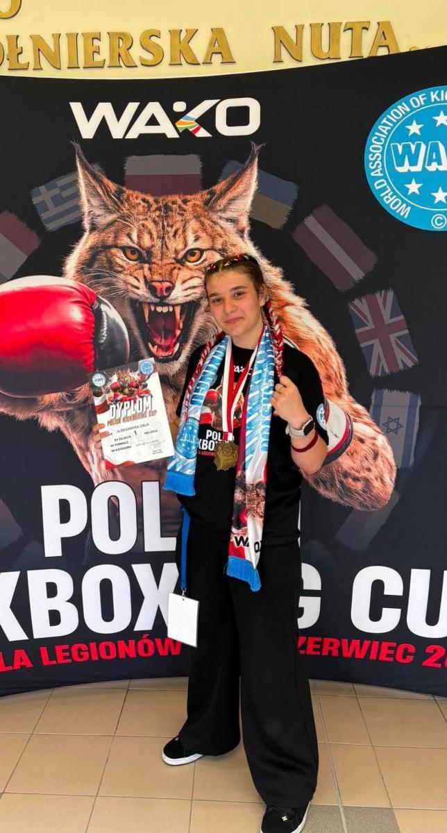 Aleksandra Sala stoi z dyplomem i medalem, w tle dziki kot, rękawica bokserska 