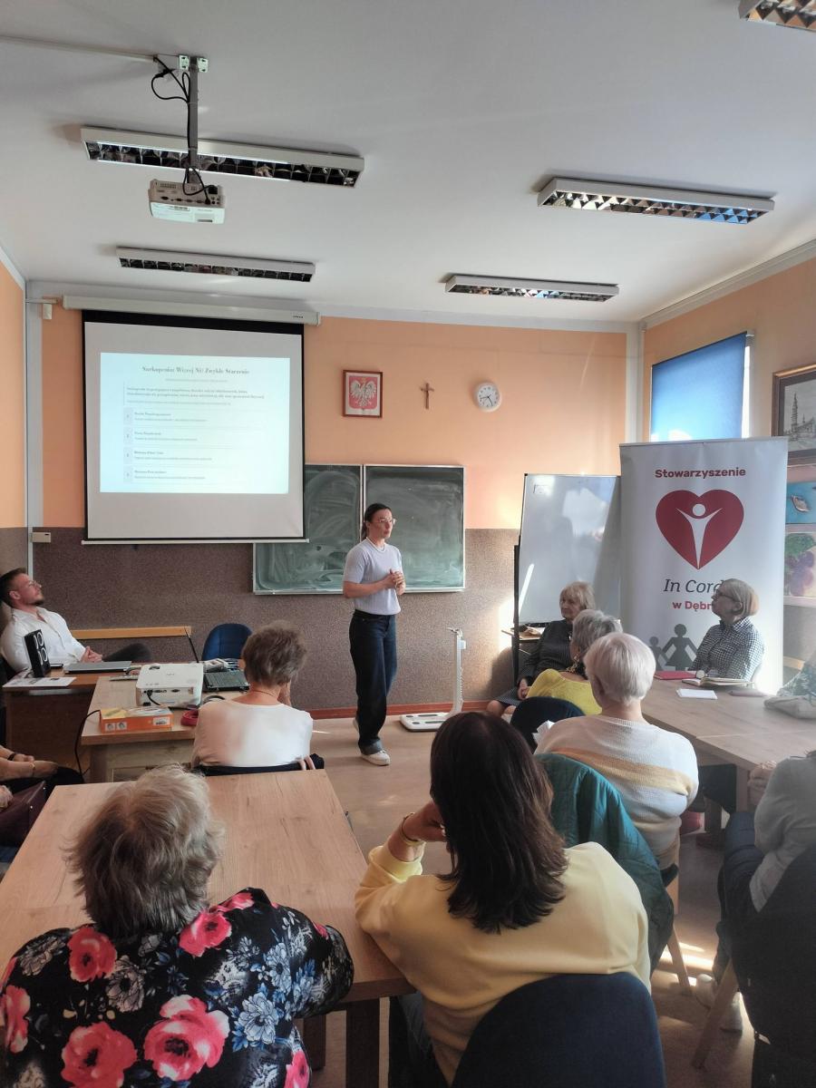 Pani prelegent stoi w sali, uczestnicy siedzą na krzesełkach 