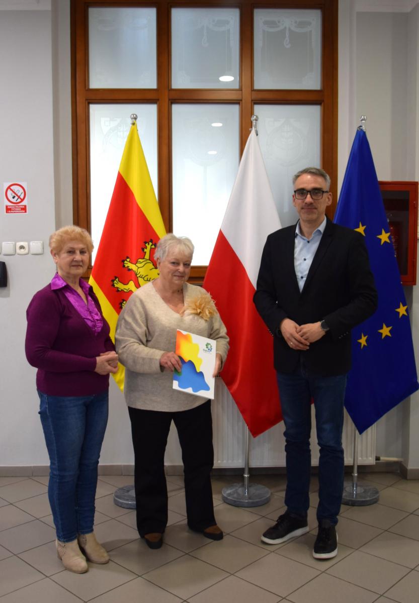 Na holu stoją, od lewej: dwie kobiety i burmistrz. W tle flaga Dębna, Polski i UE