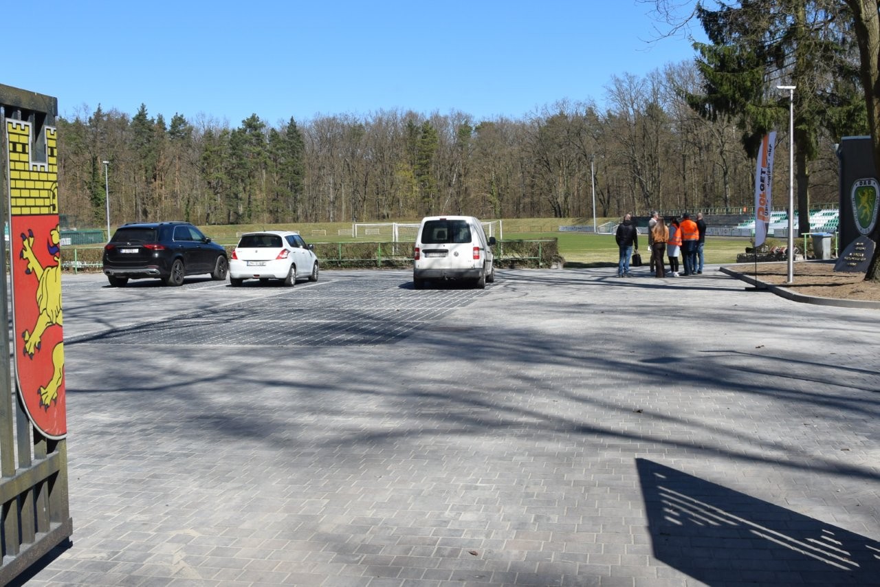 Utwardzony wjazd na stadion miejski, w tle na parkingu trzy smaochody i grupa ludzi 