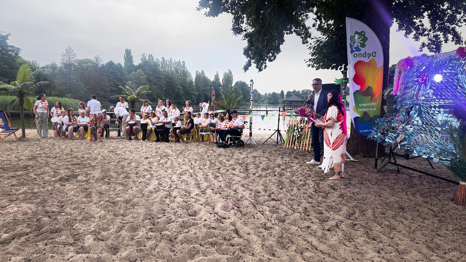 Plaża z piaskiem, na niej uczestnicy imprezy siedzą na krzesełkach, z lewej stoi burmistrz i pani kierownik 