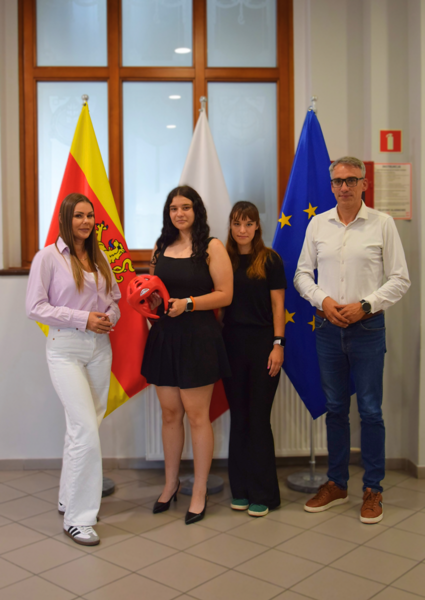 Fotografia przedstawia cztery osoby stojące wewnątrz budynku, na tle trzech flag: Gminy Dębno (ż&oacute;łto-czerwona z herbem), Polski (biało-czerwona) i Unii Europejskiej (niebieska z gwiazdami). Od lewej stoją: kobieta w jasnej koszuli i białych spodniach, młoda kobieta w czarnej sukience trzymająca czerwony kask sportowy, kolejna młoda kobieta w czarnym stroju, oraz mężczyzna w białej koszuli i jeansach. Wszyscy patrzą w obiektyw.
