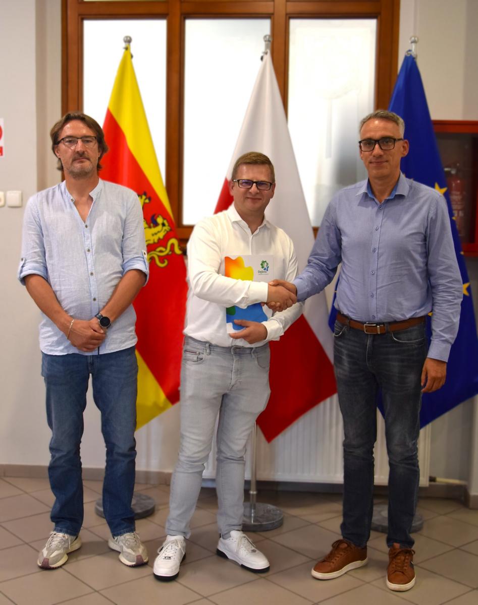 Trzej mężczyźni, stoją w Urzędzie Miejskim w Dębnie na tle flag urzędowych w kolorach ż&oacute;łtym, biało-czerwonym i niebieskim z gwiazdami unijnymi. W centralnej części mężczyzna w jasnej koszuli i jasnych spodniach trzyma dokument w teczce z logo gminy i ściska dłoń stojącego po prawej stronie Burmistrza Dębna Wojciecha Czepułkowskiego. Po lewej stronie stoi trzeci mężczyzna z rękami splecionymi z przodu. Wszyscy patrzą w obiektyw.