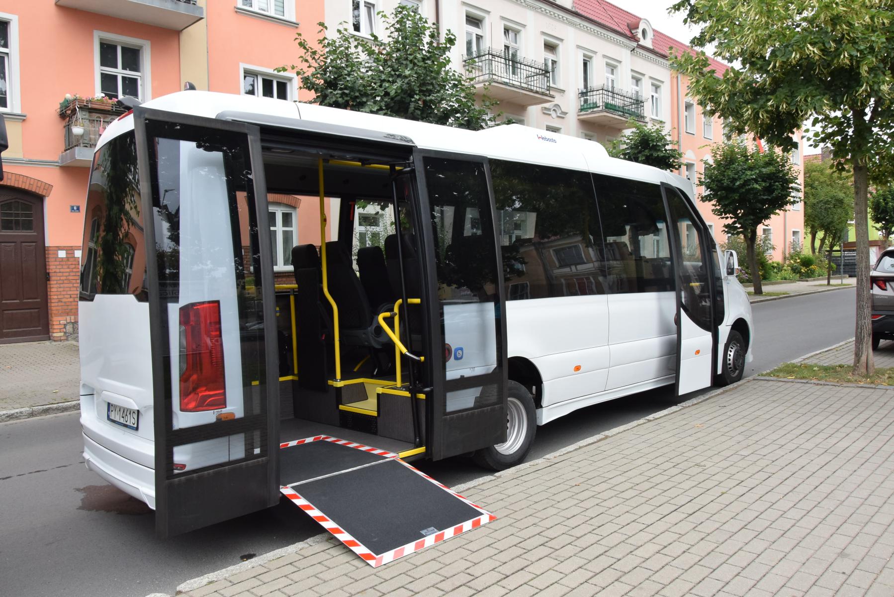 Biały autobus do przeowzy, z obnizoną rampą dla w&oacute;zk&oacute;w. Stoi na ulicy przy chodniku, w tle budynki