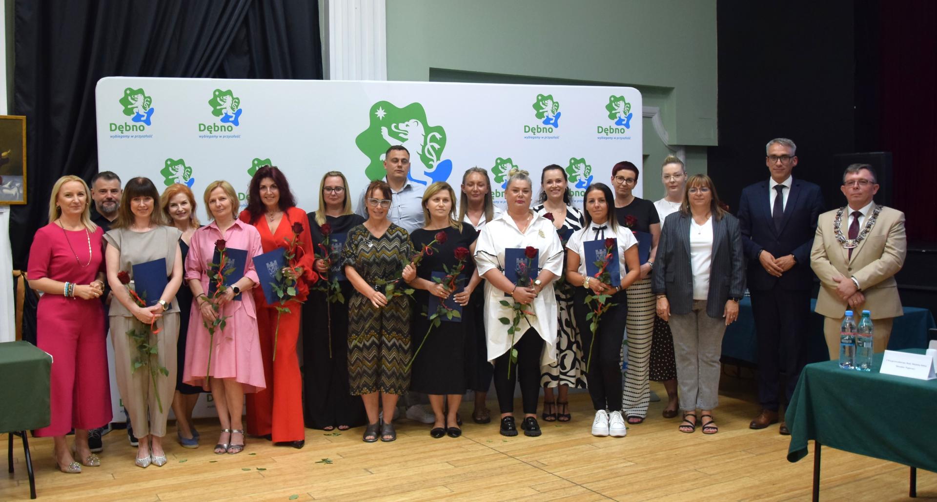 Grupa mianowanych nauczycieli w sali wraz z burmistrzem, przewodniczącm i dyrkeotrami plać&oacute;wek. W tle biała ścianka z logiem gminy Dębno.