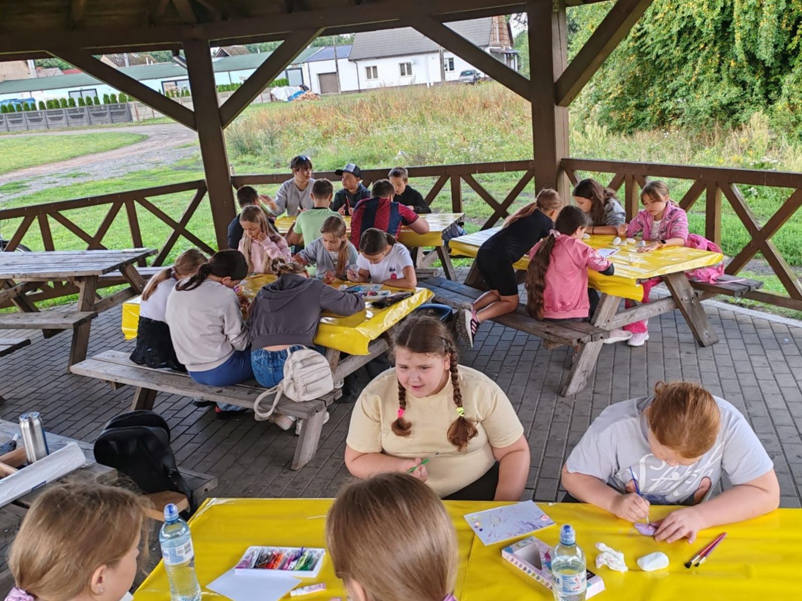 Dzieci siedzą przy stołach pod wiatą, kolorują