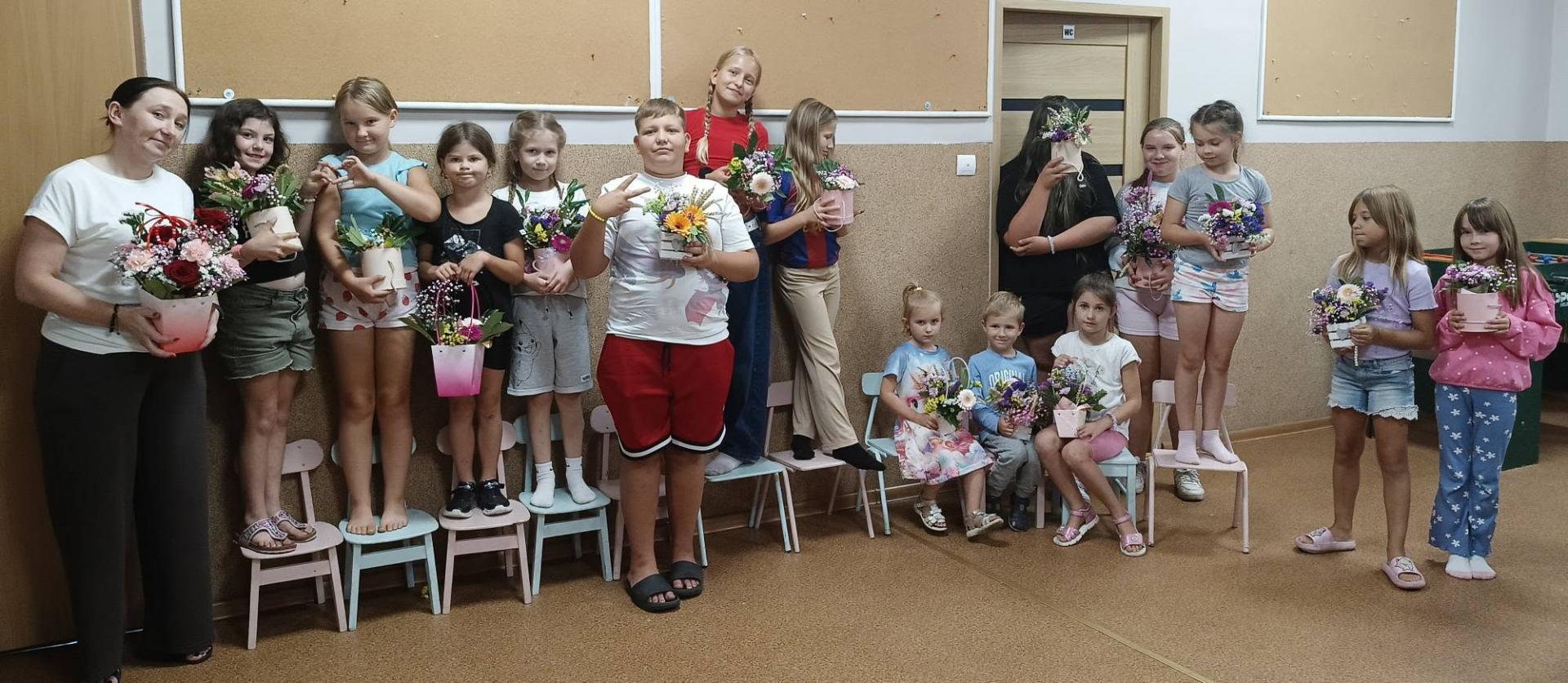 Grupa dzieci ustawiona w rzędzie w świetlicy, trzymają w rękach kwiatki w pojemnikach 