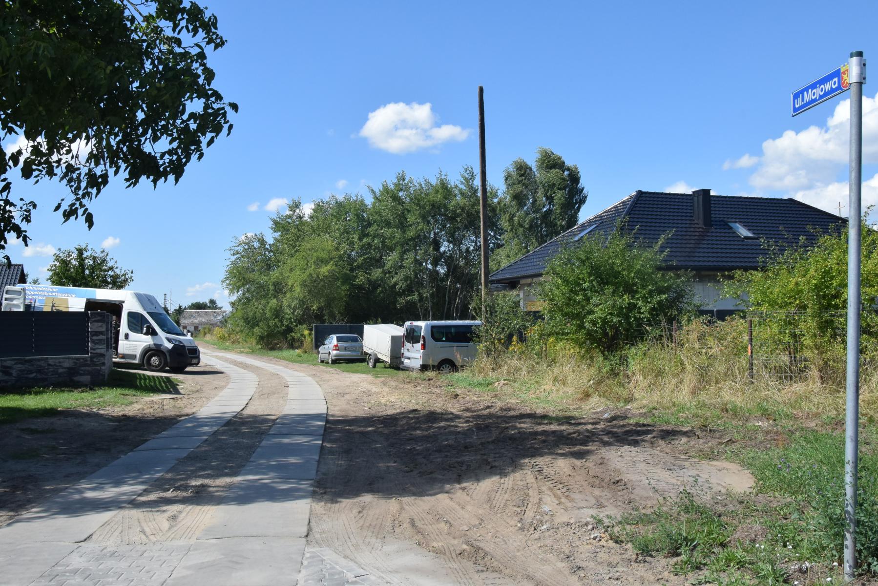 Wjazd na drogę ułozoną z dw&oacute;ch pas&oacute;w płyt, po bokach stoją busy, rosną drzewa i krzaki. 