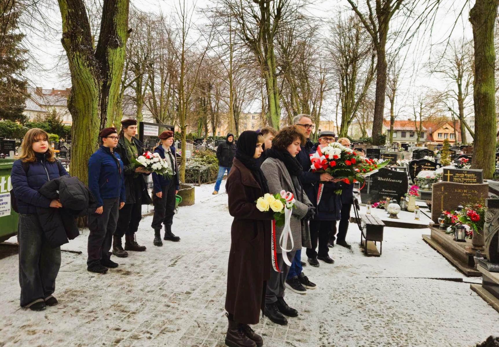Uczestnicy uroczystosci stoją na alejce cmentarnej. W tle drzewa. 