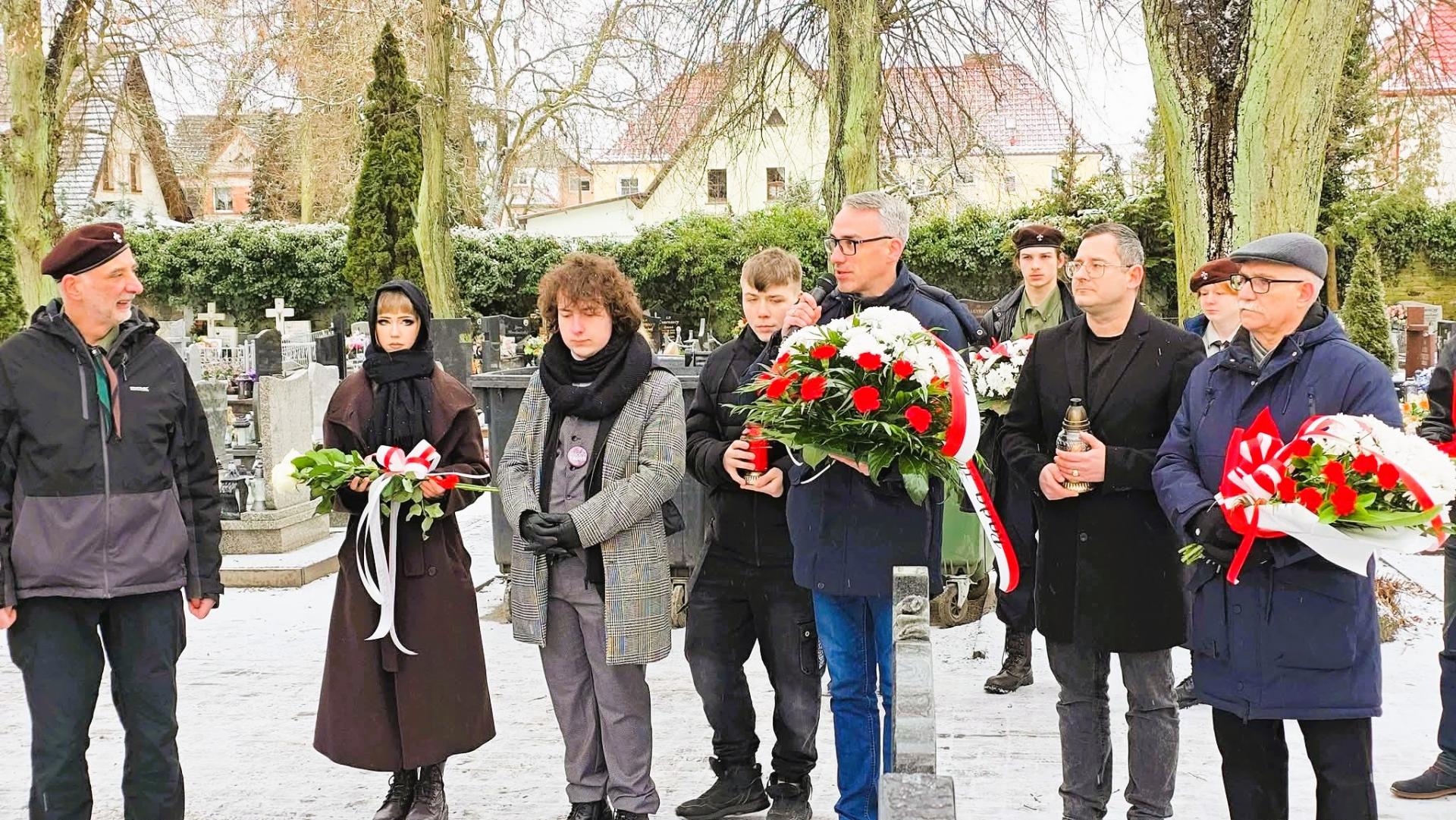 Burmistrz Dębna z biało-czerwoną wiązanką kwiat&oacute;w, przemawia, obok stoją uczestnicy młodzież, harcerze, radny