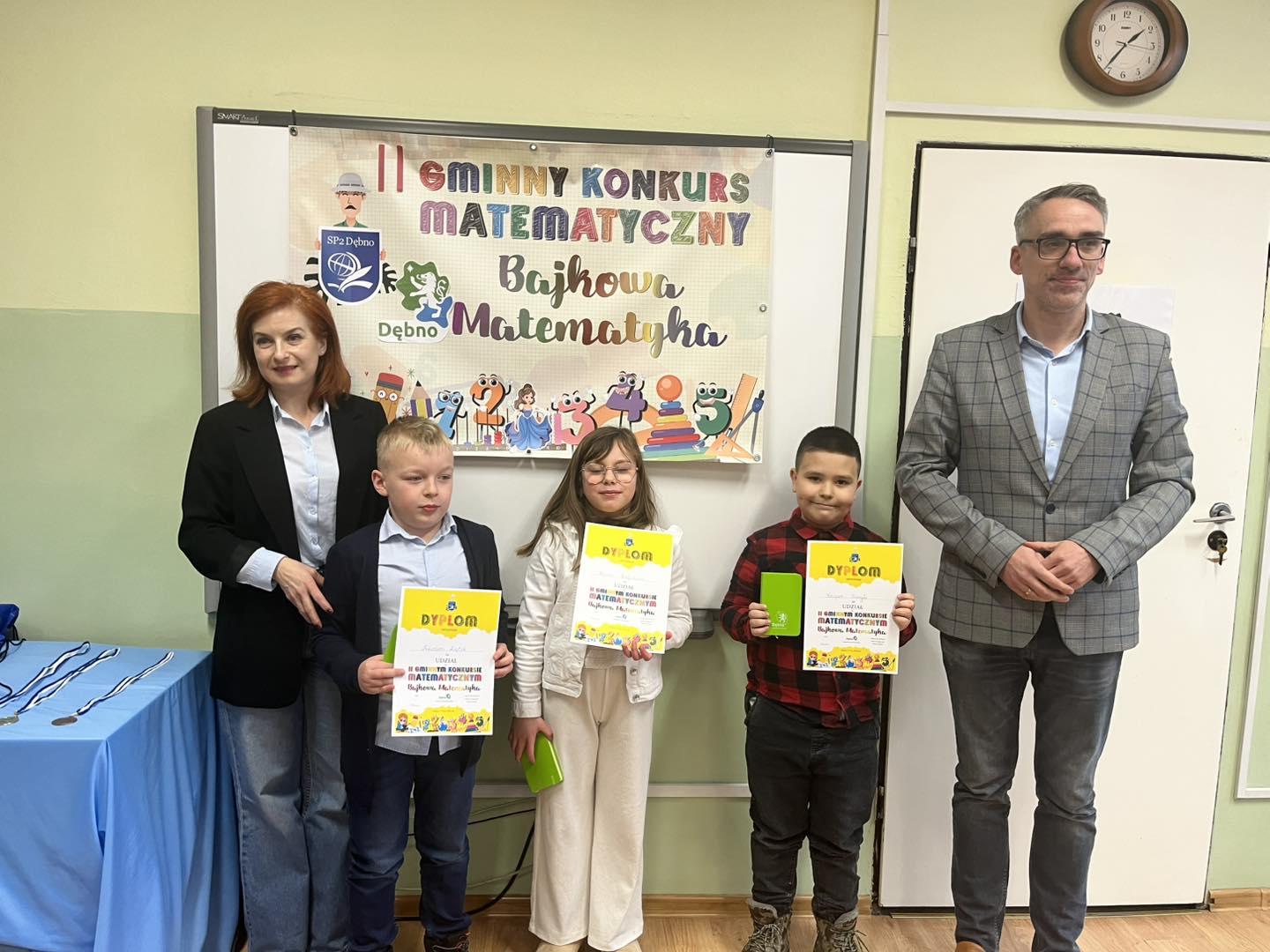 Wyr&oacute;żnieni uczniowie stoją z dyplomami w rękach, w klasie wraz z panią dyrektor (od lewej) i burmistrzem (od prawej)