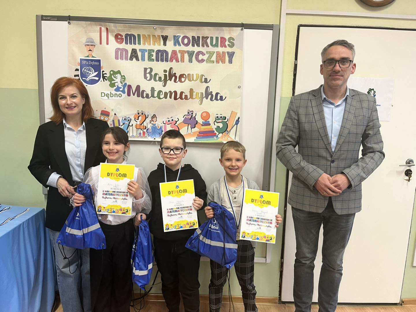 Od lewej w klasie stoja: pani dyrektor, dziewczynka, dw&oacute;ch chłopc&oacute;w trzymają w rękach dyplomy i worki, burmistrz
