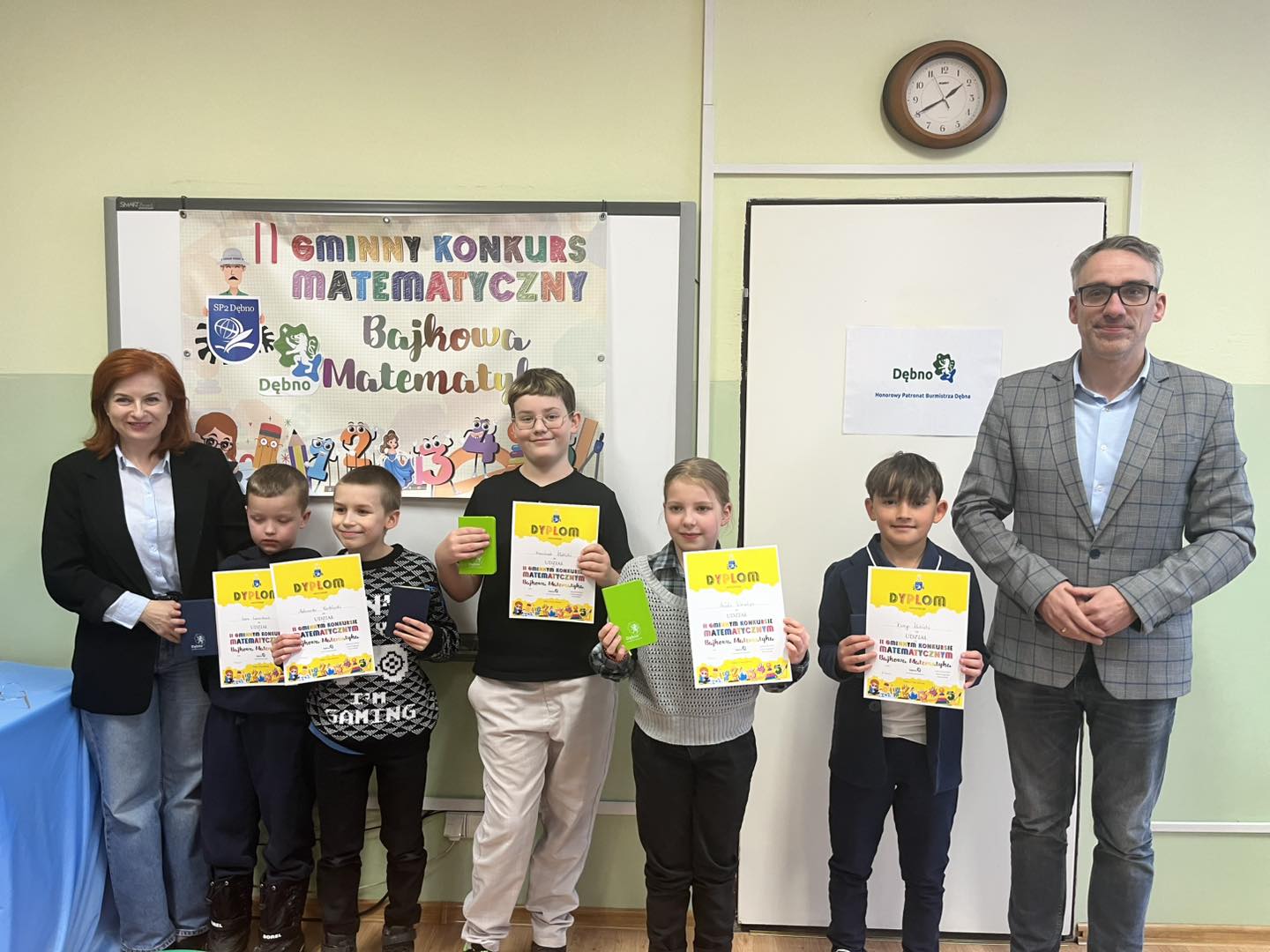 Od lewej w klasie stoją: pani dyrektor, piątka uczni&oacute;w, trzymaja dyplomy w rękach, burmistrz