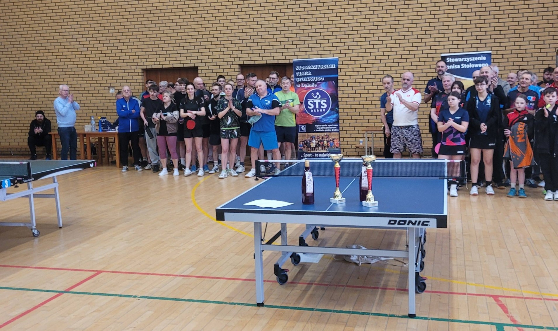 Grupa kilkudziesięciu os&oacute;b stoi na hali, przed nimi stoły do gry w tenisa