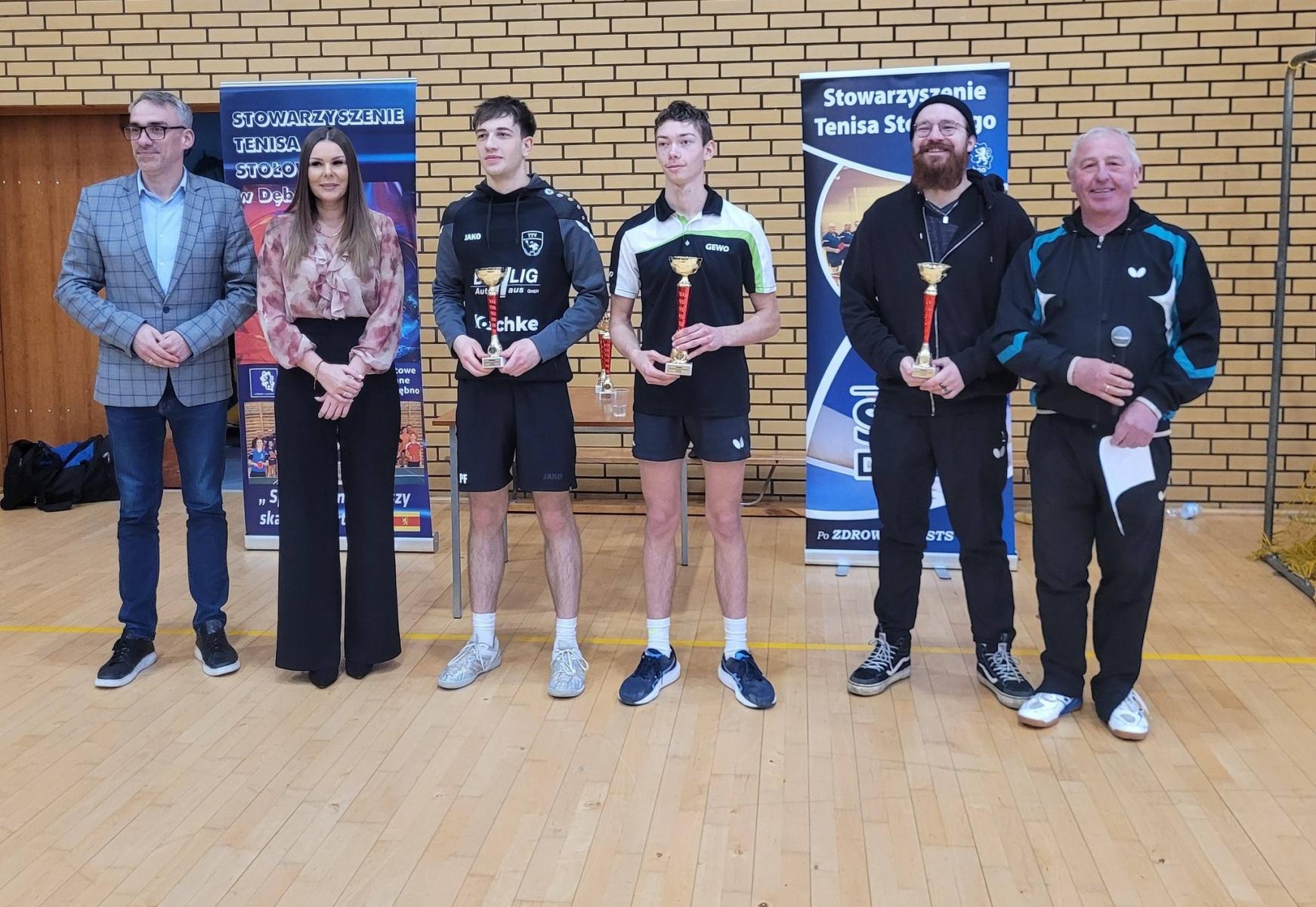 Od lewej na hali stoją: burmistrz, pani dyrektor, trzech chłopak&oacute;w z pucharami oraz prezes STS