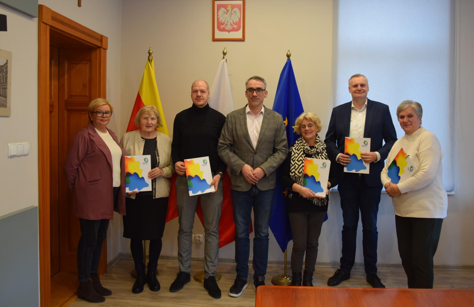 Przedstawiciele stowarzyszeń wraz z burmistrzem stoją w sali posiedzeń. w rękach trzymają teczki 