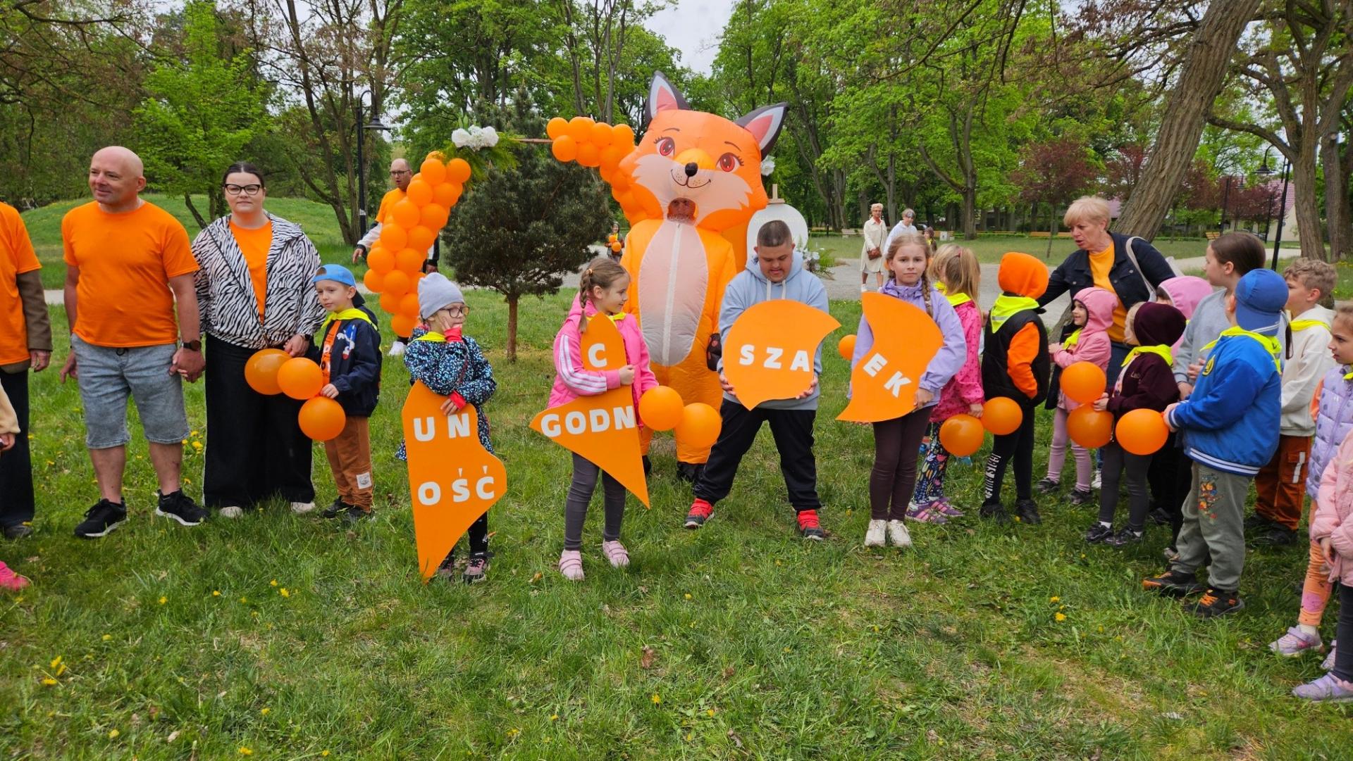 Dzieci stoją w parku, trzymja puzzle , za nimi stoi lisek 