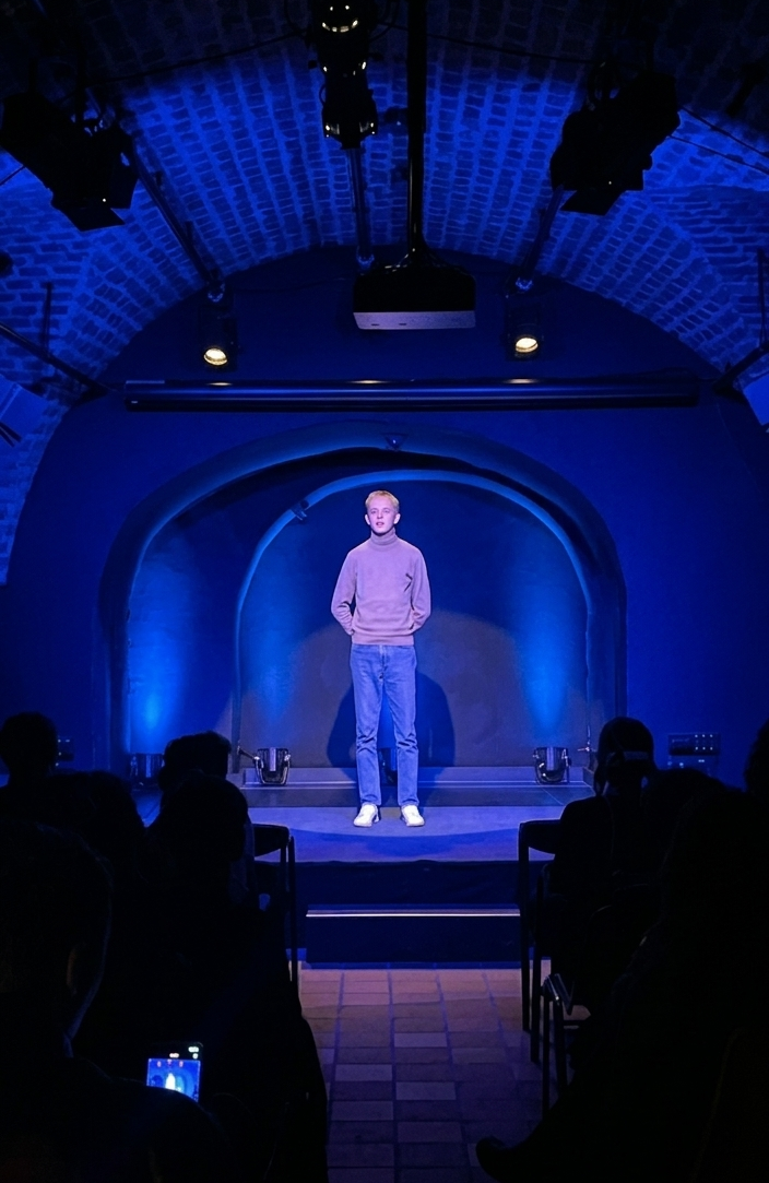 Antonii Wolski stoi na scenie, sala przyciemniona