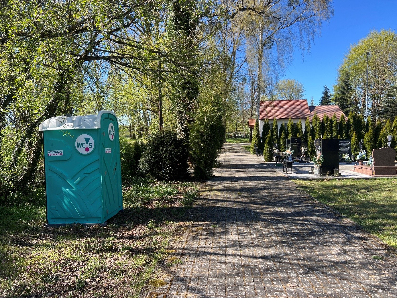 Przenośna zielona toaleta, po prawej stronie, zielone drzewa, alejka, krzewy